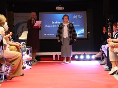 Kobieta stoi na czerwonym wybiegu w szarej, dzianinowej spódnicy, białej bluzce z ażurowym wykończeniem oraz luźnym swetrze w dużą kratę