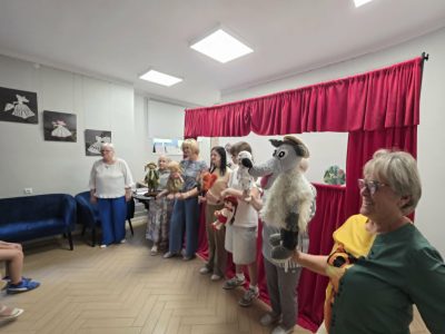 Kobiety stojące przed czerwoną kurtyną teatralną prezentują lalki i pacynki, w tym postać wilka, gajowego oraz babci. Na pierwszym planie po prawej stronie uśmiecha się kobieta w zielonej bluzce trzymająca wełnianą pacynkę wilka