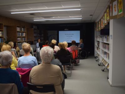 Widok z tyłu na salę biblioteczną wypełnioną siedzącymi na krzesłach uczestnikami wykładu. W głębi sali, na środku czarnej ściany, wyświetlany jest jasny slajd prezentacji z napisem „Jak odzyskać siebie i żyć bez lęku”. Po bokach ekranu siedzą prowadzące. Wokół sali ciągną się regały z książkami