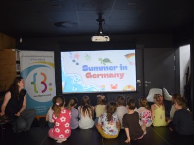 Grupa dzieci siedzi tyłem na podłodze, oglądając wyświetlany na dużym ekranie slajd z napisem „Summer in Germany” i rysunkiem żółwia morskiego. Po lewej stronie na podwyższeniu siedzi prowadząca zajęcia, obok której stoi pionowy baner reklamowy biblioteki