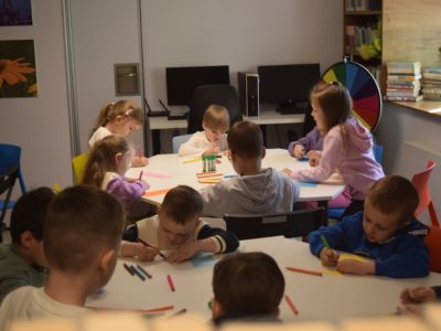 Grupa dzieci siedzi przy dużym, białym, okrągłym stole w jasnym pomieszczeniu. Dzieci rysują kredkami na kartkach papieru. W tle widoczne są stanowiska komputerowe oraz półki z książkami