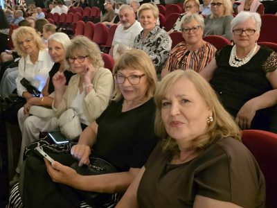 Widownia w teatrze lub kinie wypełniona publicznością siedzącą w czerwonych fotelach. Na pierwszym planie dwie uśmiechnięte kobiety patrzą w stronę obiektywu; jedna z nich trzyma w dłoni smartfon. W rzędach za nimi widać pozostałych uczestników wydarzenia, w większości starsze osoby