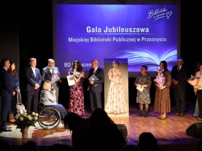 Grupa przedstawicieli instytucji stojąca na scenie po obu stronach portretu historycznego z bukietami kwiatów i upominkami podczas składania życzeń dyrekcji