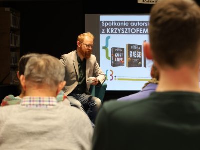 Krzysztof Piersa w jasnej marynarce siedzi w fotelu z filiżanką w dłoni i przemawia do słuchaczy. W tle na ekranie wyświetla się slajd „Spotkanie autorskie z Krzysztofem Piersą” pokazujący okładki jego książek