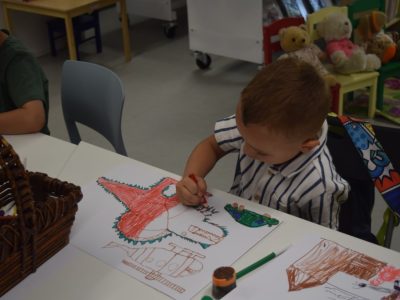 Mały chłopiec w biało-niebieskiej koszulce w paski siedzi przy białym stole, starannie kolorując rysunek czerwonego dinozaura. W tle na małych, kolorowych krzesełkach siedzą pluszowe misie