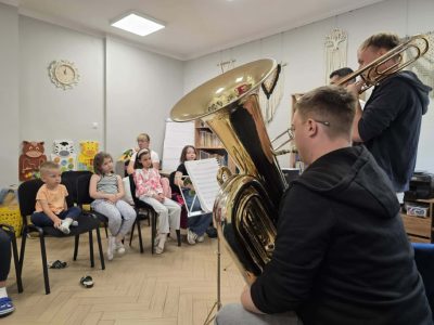 Dzieci siedzą na krzesłach i z zaciekawieniem słuchają dwóch muzyków grających na tubie i puzonie. Widok z boku na grających instrumentalistów i uśmiechniętą widownię