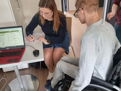 Młody mężczyzna na wózku inwalidzkim siedzi przy biurku tyłem do kadru, patrząc na ekran otwartego laptopa. Obok niego bibliotekarka w okularach i jeansowej spódnicy pochyla się nad blatem i wypełnia dokumenty papierowe
