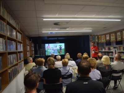 Publiczność siedząca na krzesłach w bibliotece słucha prelekcji podróżnika. Po prawej stronie w głębi stoi Daniel Kocuj w czerwonej koszulce i mówi do mikrofonu, uśmiechając się. W tle na dużym ekranie wyświetlany jest slajd ze zdjęciami z wyprawy – po lewej mężczyzna przy taczce, po prawej postać niosąca duży przedmiot na głowie pośród zieleni. Po lewej stronie kadru widoczny jest długi regał z książkami