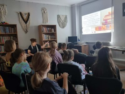 Grupa dzieci siedzi na krzesłach w sali bibliotecznej, oglądając prezentację wyświetlaną z projektora na ścianie. Prowadząca w okularach stoi przy regale z książkami i wskazuje dłonią na ekran