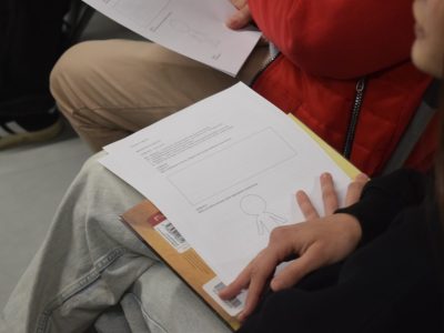 Zbliżenie na dłonie uczestnika warsztatów trzymającego kartę pracy. Na kartce widoczne są zadania dotyczące emocji oraz kontur postaci (tzw. ludzika), który służy do zaznaczania odczuć w ciele. W tle widać fragmenty ubrań innych osób biorących udział w zajęciach