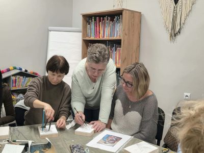 Uczestniczki warsztatów siedzą wokół stołu pokrytego folią i przygotowują szkice ikon. Prowadząca pochyla się nad stołem i pomaga jednej z kobiet w przycinaniu wzoru