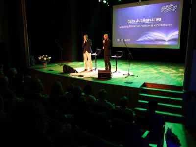 Widok z ciemnej sali na scenę oświetloną zielonym światłem, na której dwaj mężczyźni występują z mikrofonami przy hokerach. Na ekranie wyświetla się napis jubileuszowy biblioteki