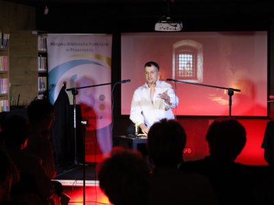Aktor w białej koszuli występuje na scenie przed mikrofonem, gestykulując dłonią. W tle widoczny jest ekran projekcyjny z ilustracją przedstawiającą okno z kratą oraz roll-up Miejskiej Biblioteki Publicznej w Przasnyszu. Na pierwszym planie widoczne są sylwetki zgromadzonej publiczności