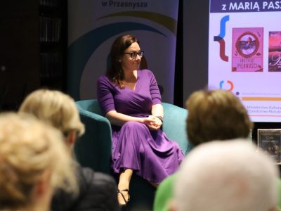 Maria Paszyńska w fioletowej sukience i okularach siedzi w turkusowym fotelu podczas spotkania autorskiego. Pisarka uśmiecha się, patrząc w stronę publiczności, której zarysy postaci widać na pierwszym planie