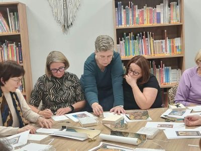 Instruktorka pochyla się nad stołem i wskazuje palcem element na kartce przed jedną z uczestniczek. Pozostałe kobiety przy stole uważnie przyglądają się i wykonują własne szkice