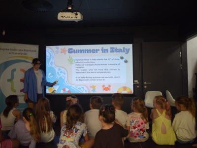 Kobieta w okularach i niebieskiej koszuli stoi przy ekranie projekcyjnym, wskazując na slajd z nagłówkiem „Summer in Italy”. Przed ekranem siedzi tyłem grupa dzieci uważnie słuchających prelekcji