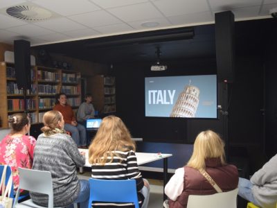 Uczestnicy Kawiarenki Językowej zgromadzeni w sali multimedialnej biblioteki patrzą na ekran, na którym wyświetla się slajd z napisem „ITALY” oraz zdjęciem Krzywej Wieży w Pizie. Przy ekranie siedzą uśmiechnięte prowadzące spotkanie