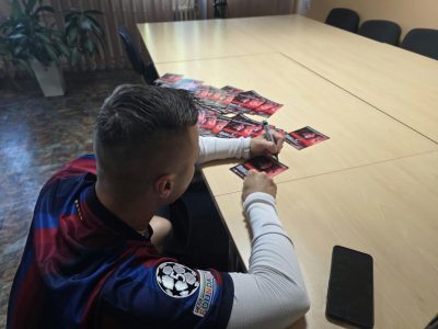 Ujęcie zza ramienia mężczyzny w koszulce FC Barcelony, który czarnym markerem składa autografy na stosie czerwonych kart promocyjnych leżących na jasnym stole