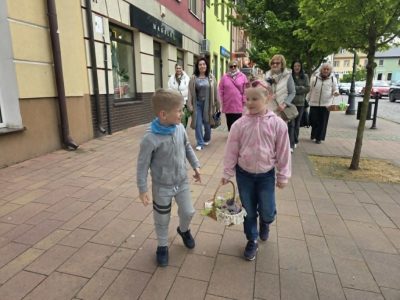 Grupa osób idzie chodnikiem wzdłuż kolorowych kamienic. Na pierwszym planie dwoje dzieci: chłopiec w szarym dresie i dziewczynka w różowej bluzie, która niesie wiklinowy koszyk. Za nimi podąża grupa dorosłych kobiet. Panuje pogodna, spacerowa atmosfera