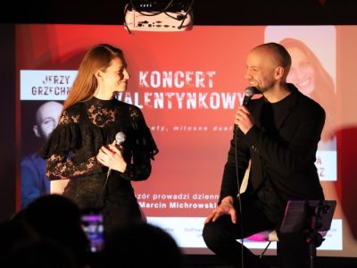 Natalia Srokocz i Jerzy Grzechnik na scenie podczas występu duetu. Obydwie osoby trzymają mikrofony i patrzą na siebie z uśmiechem. W tle wyświetlany jest czerwony slajd z napisem „Koncert Walentynkowy – duety, miłosne duety...”