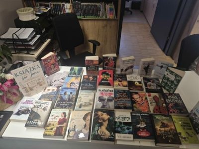 Widok z góry na biały stół w bibliotece, na którym równomiernie ułożono kilkadziesiąt książek popularnych autorów literatury obyczajowej, kryminalnej i sensacyjnej. Wśród nich wyróżnia się pionowo ustawiona tabliczka z napisem "Książka na św. Jana". W tle widoczne jest czarne biurowe krzesło obrotowe oraz regały z literaturą