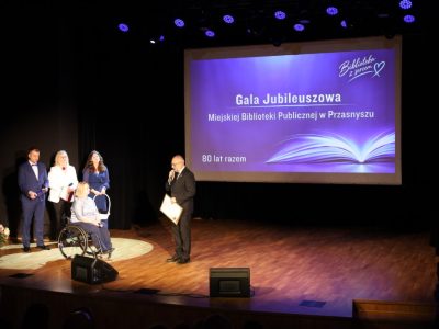 Mężczyzna w garniturze przemawia do mikrofonu, trzymając oprawiony dyplom przed gospodarzami wydarzenia. W tle znajduje się niebieski ekran z napisem „Gala Jubileuszowa Miejskiej Biblioteki Publicznej w Przasnyszu – 80 lat razem”