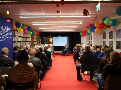 Widok od tyłu na widownię zgromadzoną w bibliotece na spotkaniu z podróżnikiem Kamilem Mąkoszą. Pośrodku sali biegnie czerwony dywan, przy regałach i na suficie wiszą kolorowe balony, a na scenie z tyłu widać prelegenta stojącego obok ekranu oraz dyrektor biblioteki