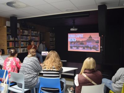 Uczestnicy Kawiarenki Językowej siedzą tyłem na krzesłach przy stolikach w sali bibliotecznej, oglądając prezentację wyświetlaną na ekranie. Na slajdzie widnieje napis „Roma Caput Mundi” oraz zdjęcie Rzymu z widokiem na Bazylikę św. Piotra. Z boku stoją prelegentki prowadzące spotkanie