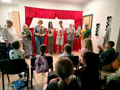 Grupa seniorek stoi na scenie za czerwoną kurtyną teatrzyku kukiełkowego, prezentując dzieciom trzymane w rękach pacynki przedstawiające postaci z bajki o Czerwonym Kapturku. Widownia siedząca tyłem na krzesłach obserwuje występ