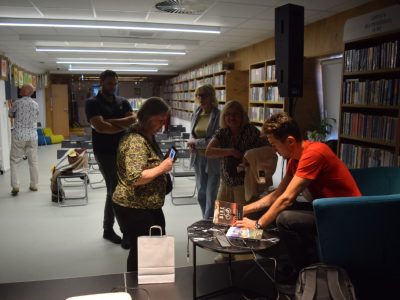 Daniel Kocuj w czerwonej koszulce siedzi w turkusowym fotelu i podpisuje książki przy małym, okrągłym stoliku w bibliotece. Przed nim stoi grupa czytelników – na pierwszym planie kobieta w kwiecistej bluzce przegląda coś w telefonie, obok stoi mężczyzna z ramionami splecionymi na piersi, a w tle czekają kolejne dwie kobiety. W głębi sali widać rzędy pustych krzeseł i regały z książkami