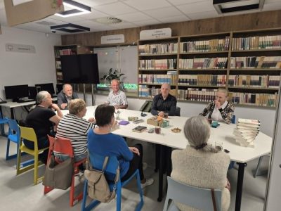 Grupa osób siedzi przy dużym, białym stole w nowoczesnej sali bibliotecznej, rozmawiając i pijąc napoje z szklanek. Na stole leżą książki, okulary oraz drobne przekąski. W tle widać wysokie, drewniane regały wypełnione książkami z oznaczeniem sekcji lektur dla młodzieży, a po lewej stronie stanowiska komputerowe z monitorami