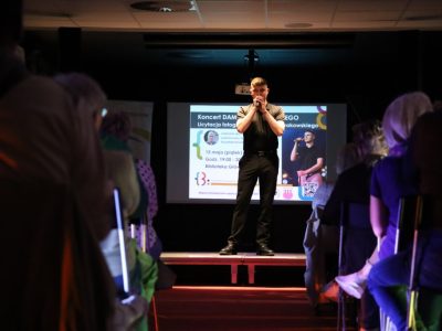Koncert Damiana Kowalskiego w bibliotece. Młody mężczyzna w czarnej koszulce polo i czarnych spodniach śpiewa do mikrofonu na podwyższeniu sceny. Za nim wyświetlany jest slajd informacyjny o koncercie i licytacji charytatywnej z datą 15 maja. Widownia obserwuje występ w przyciemnionej sali