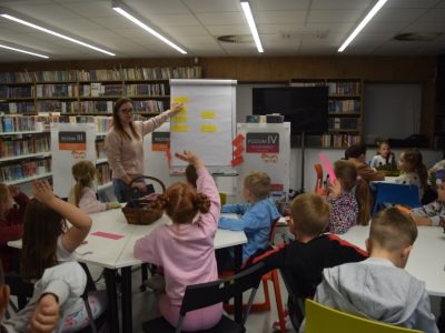 Grupa dzieci siedzi przy stołach w jasnej bibliotece, słuchając kobiety stojącej przy tablicy flipchart. Prowadząca wskazuje ręką na naklejone na tablicy żółte kartki z działaniami, a niektóre dzieci podnoszą ręce, zgłaszając się do odpowiedzi