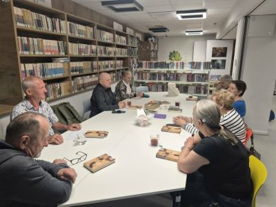 Widok z boku na uczestników dyskusji siedzących przy białym stole konferencyjnym w bibliotece. Przed kilkoma osobami leżą egzemplarze tej samej książki z widoczną ilustracją na okładce. Wszyscy są zwróceni ku sobie, zaangażowani w rozmowę. Tło stanowią regały z książkami oraz fragment strefy dziecięcej z zielonym smokiem na ścianie