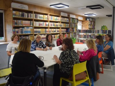 Grupa ośmiu kobiet siedzi przy dużym, białym stole w nowoczesnej sali bibliotecznej. Kobiety rozmawiają, piją kawę i herbatę. W tle widoczne są wysokie, drewniane regały wypełnione książkami oraz kolorowe krzesła. Jedna z uczestniczek porusza się na wózku