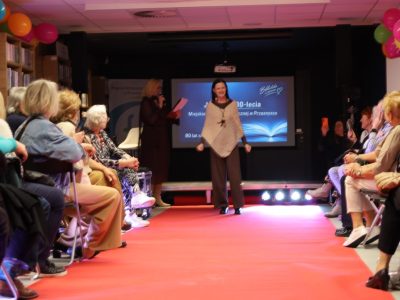 Uśmiechnięta modelka w luźnym, dzianinowym ponczo w odcieniach beżu i brązu oraz szerokich spodniach pozuje na środku czerwonego dywanu w bibliotece