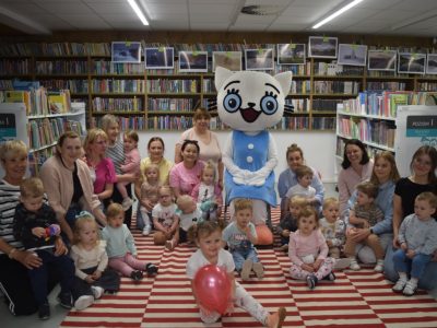 Pamiątkowe zdjęcie grupowe uczestników spotkania w bibliotece. Ponad piętnaścioro małych dzieci oraz kilka dorosłych kobiet siedzi i klęczy na pasiastym dywanie. W samym centrum grupy stoi maskotka Kici Koci w niebieskiej sukience. W tle znajdują się wysokie regały wypełnione książkami, nad którymi zawieszono galerię zdjęć. Na pierwszym planie dziewczynka trzyma różowy balon