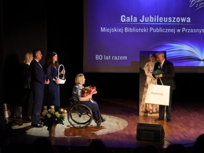 Mężczyzna w garniturze stoi na scenie z papierową torbą „Mazowsze” oraz teczką i przemawia do mikrofonu w stronę dyrektor biblioteki.