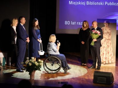 Mężczyzna w garniturze trzymający bukiet kwiatów oraz stojąca obok niego kobieta w czarnej sukience składają życzenia dyrektor biblioteki