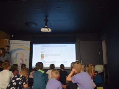 Grupa dzieci siedzi tyłem na podłodze w zaciemnionej sali, oglądając wyświetlaną na dużym ekranie stronę internetową bibliotecznej wyszukiwarki książek. Obok ekranu stoi baner reklamowy, a z boku przygląda się im opiekunka