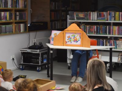 Bibliotekarka siedząca przy białym stoliku w otoczeniu regałów z książkami przedstawia dzieciom bajkę za pomocą drewnianego teatrzyku Kamishibai. Widzowie siedzą tyłem na dywanie