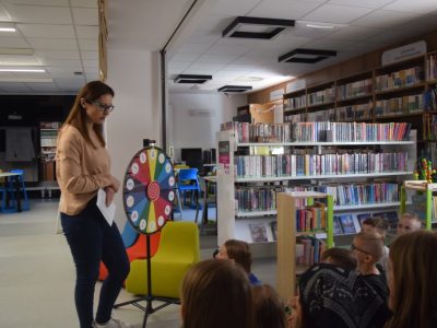 Prowadząca warsztaty kobieta stoi w jasnym wnętrzu biblioteki i trzyma w rękach kartkę. Obok niej umieszczone jest duże, kolorowe koło fortuny z polami oznaczonymi cyframi. Przed kobietą siedzi grupa dzieci słuchających instrukcji. W tle widać regały z książkam