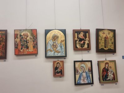 Osiem ikona sakralnych przedstawiających postacie religijne w ozdobnych szatach, zawieszonych na linkach na białej ścianie galerii w dwóch rzędach