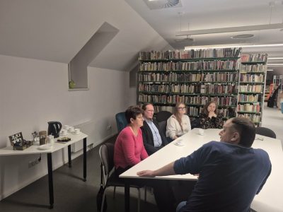 Widok z boku na uczestników spotkania siedzących przy białym stole w bibliotece. Na pierwszym planie po prawej stronie tyłem do obiektywu siedzi mężczyzna w niebieskiej bluzie. Naprzeciwko niego, wzdłuż stołu, siedzą cztery osoby patrzace w jego kierunku. W tle po lewej stronie przygotowano stolik kawowy z czajnikiem i filiżankami