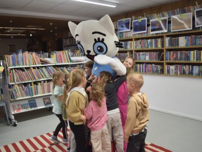 Grupa uśmiechniętych przedszkolaków tuli się do stojącej na środku sali bibliotecznej maskotki Kici Koci. Dzieci otaczają postać ciasnym kręgiem, okazując radość. W tle widoczne są regały z książkami oraz wiszące pod sufitem zdjęcia w ramkach