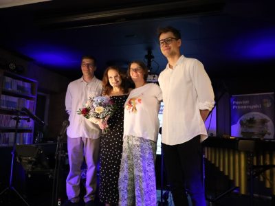 Czworo uśmiechniętych ludzi pozuje do zdjęcia na scenie po zakończonym występie. Wśród nich jest wokalistka Joanna Bejm, trzymająca duży bukiet kolorowych kwiatów i ubrana w czarną sukienkę w białe grochy. Obok niej stoją dwaj muzycy w białych koszulach oraz uczestniczka. Scena oświetlona jest niebieskim światłem, a w tle widać instrumenty muzyczne oraz baner z napisem Powiat Przasnyski