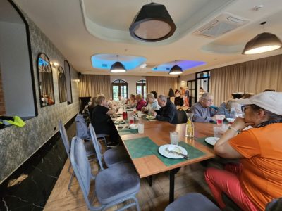 Uczestnicy wydarzenia siedzą przy długim, drewnianym stole w nowocześnie urządzonej jadalni lub restauracji z ozdobnymi sufitami o niebieskim podświetleniu. Ludzie jedzą posiłek, rozmawiają i piją napoje. Na pierwszym planie po prawej stronie siedzi kobieta w pomarańczowej koszulce i jasnej czapce z daszkiem