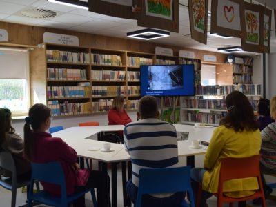 Grupa osób siedzi przy dużym, stole w bibliotece. Przed nimi znajduje się włączony monitor prezentujący zdjęcie remontu budynku. W tle widoczne są wysokie regały wypełnione książkami z oznaczeniami działów