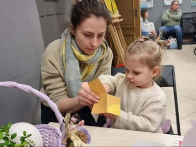 Na pierwszym planie widać kobietę, która pomaga małej dziewczynce robić wielkanocną pracę plastyczną. Obie siedzą przy stoliku, naprzeciw nich stoi wiklinowy kosz z ozdobą. Kobieta najprawdopodobniej jest mamą dziewczynki