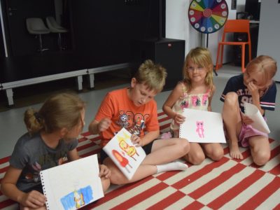 Czworo dzieci siedzi na dywanie w biało-czerwone pasy i pokazuje do aparatu swoje rysunki przedstawiające kolorowe postacie, w tym różowego misia i postać w płomieniach